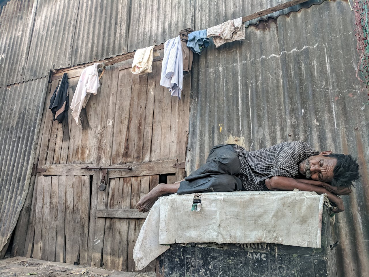 services-04 An elderly man peacefully sleeps against a rundown wooden wall with hanging clothes.